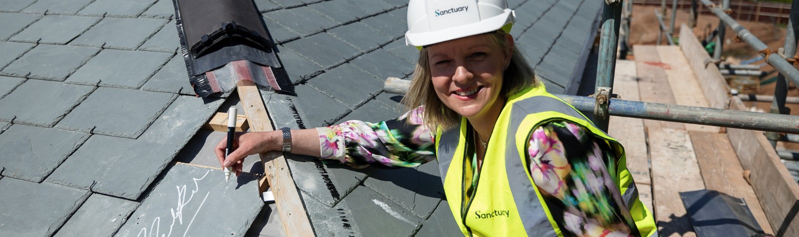 A female with blonde hair wearing a hard hat and high-vis on top of the roof of the care home signing a tile with a white pen